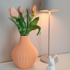 May include: A peach-colored, ribbed vase holds faux tulips with green leaves. A small, white bunny figurine sits on a white surface. A gray lamp with a matching lampshade is in the background.