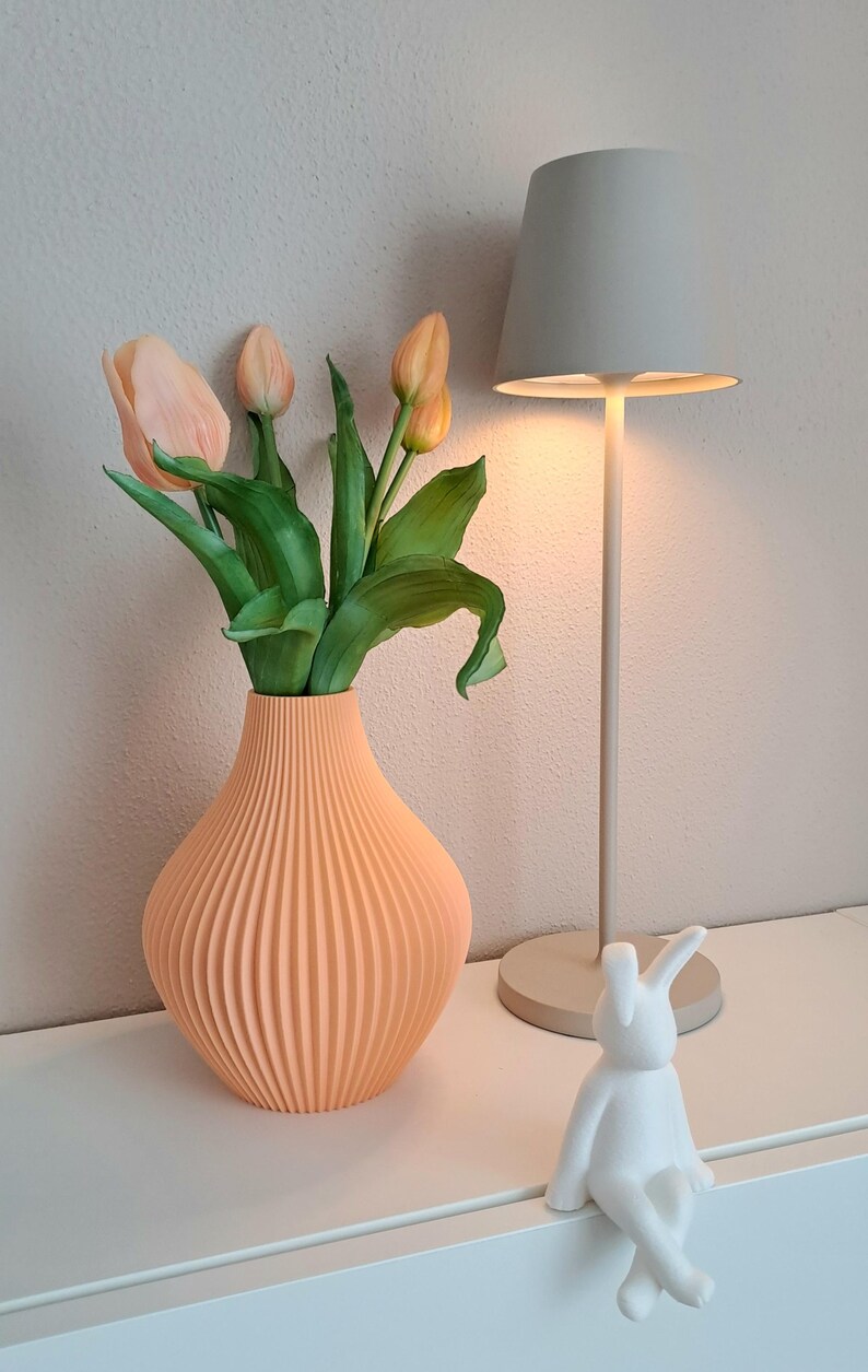 May include: A peach-colored, ribbed vase holds faux tulips with green leaves. A small, white bunny figurine sits on a white surface. A gray lamp with a matching lampshade is in the background.