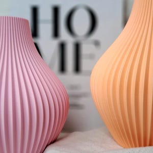 May include: Two decorative vases with a ribbed design. One vase is a soft pink, the other is a light orange. The vases are set against a blurred background with the word "HOME" visible. The vases are on a white cloth.