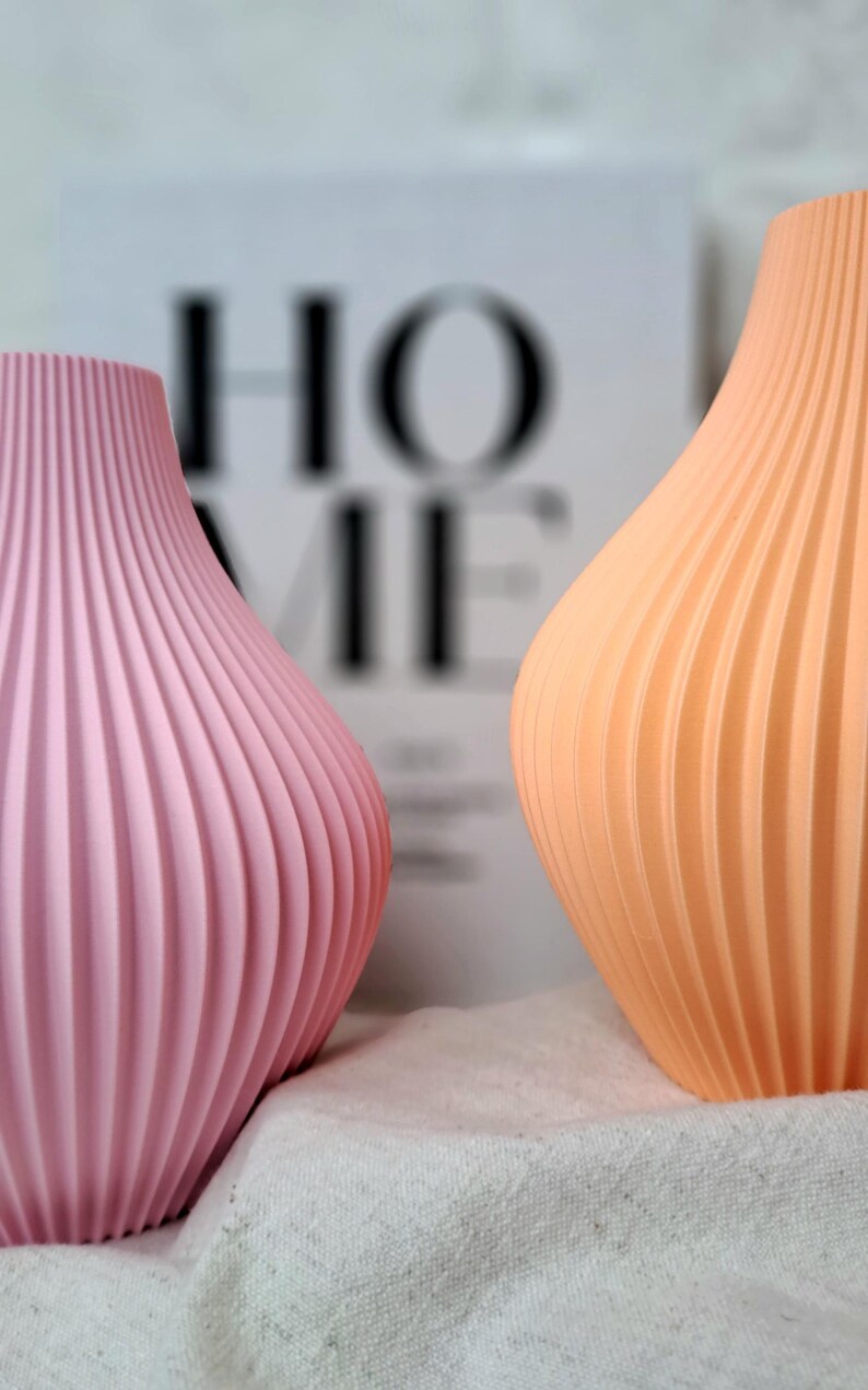 May include: Two decorative vases with a ribbed design. One vase is a soft pink, the other is a light orange. The vases are set against a blurred background with the word "HOME" visible. The vases are on a white cloth.