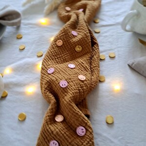 May include: A table setting featuring a mustard-coloured fabric runner decorated with pink smiley face buttons. Plates, napkins, and mugs are arranged on the table. Gold confetti and fairy lights provide a festive atmosphere.
