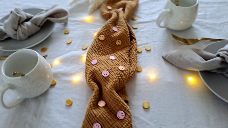May include: A table setting featuring a mustard-coloured fabric runner decorated with pink smiley face buttons. Plates, napkins, and mugs are arranged on the table. Gold confetti and fairy lights provide a festive atmosphere.