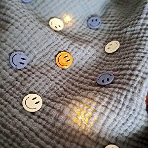 May include: Various coloured smiley face buttons, including yellow, white, and blue, scattered on a textured, grey fabric. The buttons have a simple, cheerful design, and the fabric has a crinkled appearance.