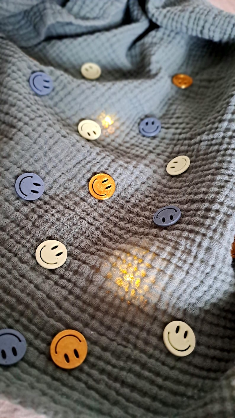 May include: Various coloured smiley face buttons, including yellow, white, and blue, scattered on a textured, grey fabric. The buttons have a simple, cheerful design, and the fabric has a crinkled appearance.