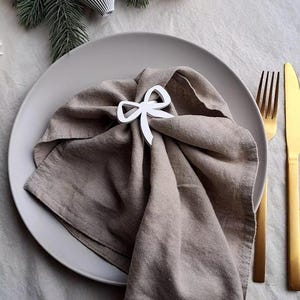 Puede incluir: Una servilleta de lino de tono neutro está doblada sobre un plato gris, asegurada con un servilletero blanco en forma de lazo. Tenedor y cuchillo dorados a la derecha. Adornos de papel y ramas de pino en el fondo.