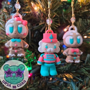 May include: Three Christmas tree ornaments in the shape of gingerbread people and a nutcracker. The ornaments are brown, white, pink, and teal. They are hanging from gold chains with clear beads. The ornaments are hanging on a Christmas tree.