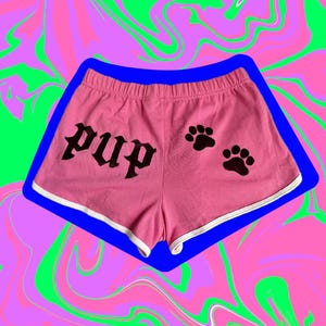 Peut inclure: Short rose avec une bordure blanche et le mot "pup" en lettres gothiques noires. Deux empreintes de pattes noires sont imprimées sur le côté droit du short. Le short est posé sur un fond coloré tourbillonnant.