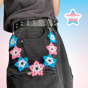 Peut inclure: Une ceinture en cuir noir avec des œillets argentés et une chaîne d'étoiles perlées. La chaîne d'étoiles est ornée de perles roses, blanches et bleues, et est fixée à la ceinture par des fermoirs argentés. La ceinture est portée avec un jean en denim noir.