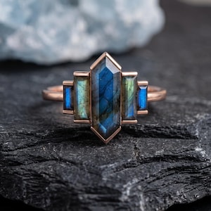 May include: A rose gold ring featuring a central hexagonal labradorite stone flanked by four smaller rectangular baguette-cut labradorite stones. The stones display iridescent blue and green flashes set within a polished metal band.
