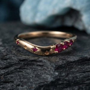 May include: A curved gold ring featuring three round red rubies set on one side and small star-shaped engravings along the band. The metal has a polished finish, and the design follows a gentle wave shape, highlighting the vibrant red gemstones.