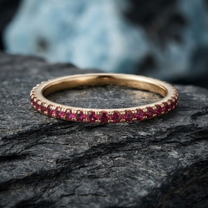 May include: A thin gold band ring featuring a continuous row of small, round, vibrant red gemstones set along the entire outer circumference. The metal has a polished finish, and the stones are uniform in size, creating a consistent circular design.