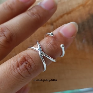 Puede incluir: Un anillo plateado con un diseño único se muestra en un dedo. El anillo tiene un elemento en forma de Y y dos pequeños acentos redondos. El anillo está en un dedo con una uña natural. El fondo es una superficie de madera.