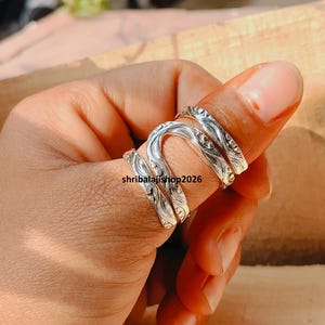 May include: A silver ring with a detailed, swirling pattern is worn on a finger. The ring wraps around the finger with a textured design. The text "shribalajishop2026" is visible on the ring.