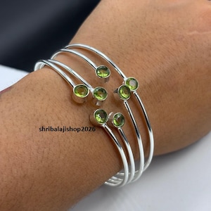 May include: A set of three silver-toned open cuff bracelets, each featuring two small, round, faceted lime-green stones set at the ends. The text shribalajishop2026 is visible on the surface of the bracelets. The design is simple and metallic.