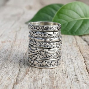 May include: A stack of silver rings with intricate floral and leaf patterns. The rings are stacked on a wooden surface, with a blurred green leaf in the background. Each ring features a unique design, showcasing detailed craftsmanship.
