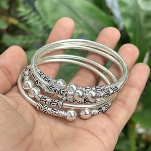 Op de afbeelding: Een set zilveren armbanden met decoratieve kralenaccenten. De armbanden zijn rond en glad, met een gepolijste afwerking. De decoratieve secties hebben ingewikkelde patronen en kleine zilveren kralen. De armbanden zijn op elkaar gestapeld.