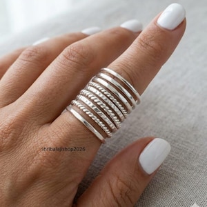 Può includere: Una collezione di anelli d'argento di vari design, tra cui fasce lisce e motivi a corda, sono impilati su un dito. Gli anelli sono mostrati su una mano con smalto bianco. Il nome del negozio Etsy, shribalajishop2026, è visibile.