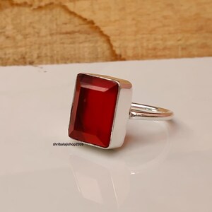 May include: A silver ring featuring a large, rectangular, faceted red gemstone set in a smooth metal bezel. The band is thin and polished silver. The text "shribalajishop2026" is printed in small black letters near the base of the ring setting.