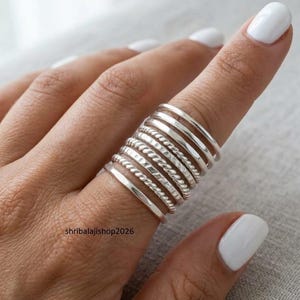 Könnte beinhalten: Eine Sammlung silberner Ringe in verschiedenen Designs, darunter glatte Bänder, gedrehte Seiltexturen und gehämmertes Finish. Die Ringe sind auf einem Finger gestapelt und zeigen die verschiedenen Stile und Texturen. Das Bild ist gut beleuchtet und hebt den metallischen Glanz des Schmucks hervor.