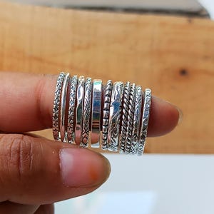 May include: A collection of silver rings with various textures and patterns, including braided, beaded, and etched designs. The rings are stacked on a finger, showcasing the different styles and widths. The rings are made of a shiny, metallic material.