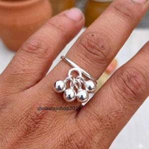 May include: A silver ring featuring five small, spherical, dangling charms. The ring is worn on a finger, and the design includes the text "shribalajishop2026". The ring is made of a polished, reflective metal.