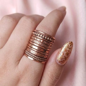 Puede incluir: Una pila de siete anillos de metal en tono cobre usados en un dedo. Los anillos presentan varias texturas, incluyendo bandas lisas, patrones de cuerda trenzada y diseños geométricos grabados. El metal tiene un acabado metálico marrón rojizo.