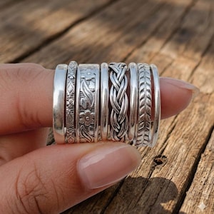 May include: A selection of silver rings featuring diverse designs, such as floral, braided, and patterned bands. The rings are presented on a wooden surface, highlighting their intricate details and textures. The rings have a mix of smooth and textured surfaces.