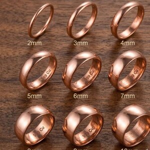 May include: A collection of copper-colored rings, each with a different width, ranging from 2mm to 10mm. The rings are displayed on a wooden surface. Each ring is marked with its size and the text "999 Cu".