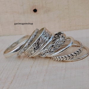 Puede incluir: Un conjunto de anillos de plata con varios diseños, incluyendo bandas florales, trenzadas y estampadas. Los anillos se exhiben sobre una superficie de madera clara. Los anillos son de diferentes anchos y estilos, mostrando una variedad de texturas y detalles.