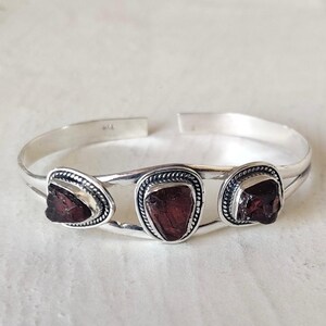 Op de afbeelding: Een zilveren manchetarmband met drie ruwe, dieprode granaatstenen gezet in gepolijst metaal. Elke steen is omlijst door een decoratieve gedraaide touwrand. De armband heeft een open ontwerp en een gladde, reflecterende afwerking op de metalen band.