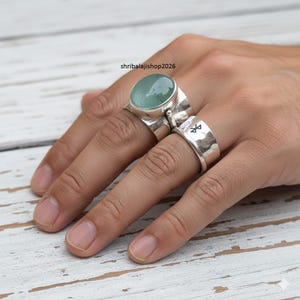 Puede incluir: Se muestran dos anillos de plata en una mano. Un anillo presenta una piedra preciosa grande, ovalada y de color verde claro. El otro es una banda gruesa con un símbolo. Los anillos están sobre una superficie de madera blanca.