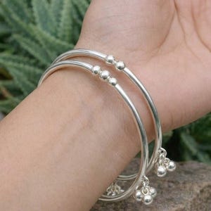 Könnte beinhalten: Zwei silberne Armreifen mit kleinen, hängenden Glockenanhängern. Die Armreifen haben eine glatte, polierte Oberfläche und sind mit kleinen, runden Perlen verziert. Der Schmuck wird an einem Handgelenk mit einem verschwommenen grünen Hintergrund gezeigt.