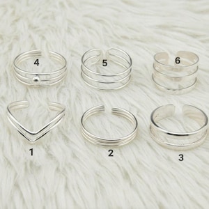 Op de afbeelding: Een collectie van zes zilverkleurige teenringen. Ontwerpen omvatten een V-vorm, meerdere banden en een ring met een kleine kraal. De ringen worden getoond op een witte, zachte ondergrond.