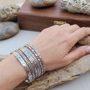 May include: A set of silver bangles with detailed embossed patterns, including floral and geometric designs. The bangles are stacked on a wrist, displaying the variety of textures and styles. The jewellery is presented on a wooden surface, with natural elements in the background.