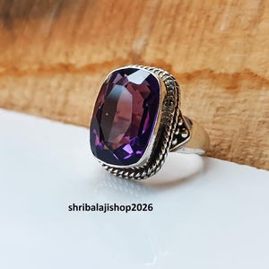 May include: A silver ring featuring a large, rectangular, faceted amethyst gemstone. The amethyst is a deep purple color and is set in a decorative silver band with a rope-like texture. The text "shribalajishop2026" is visible.