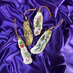Peut inclure: Quatre décorations de Noël en coquilles d'huîtres avec des bords dorés et des boucles de suspension en cuir. Deux ont des motifs floraux peints, un a un sucre d'orge et une étoile, et un est doré. Sur une surface de satin violet.