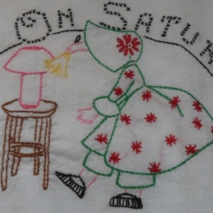 May include: Embroidered artwork on white fabric. The design features a figure in a bonnet and dress, dusting a lamp on a small stool. The text "DUST ON SATURDAY" arches above the scene, all in black thread. The figure's dress has red star accents.