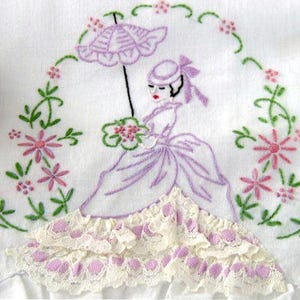 May include: A vintage embroidered white linen tea towel with a purple floral design. The design features a woman holding an umbrella and a bouquet of flowers. The towel has a lace trim.