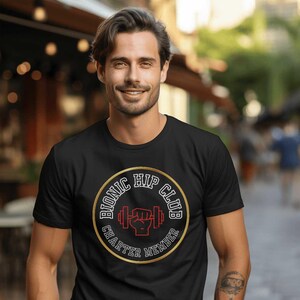 Puede incluir: Camiseta negra con un diseño gráfico circular dorado y rojo. El diseño muestra el texto "BIONIC HIP CLUB" y "CHARTER MEMBER". Un puño rojo agarrando una barra con pesas está en el centro.