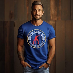 May include: A royal blue t-shirt with a gold-bordered circular graphic that reads "Bionic Knee Club Charter Member." The graphic features a red silhouette of a person playing basketball. The shirt is a crew neck.