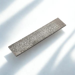 May include: A rectangular, patterned metal object with a dark border. The surface features a repeating circular design in shades of gray and silver. The object is set against a white background with shadows.