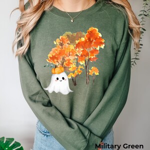 Könnte beinhalten: Ein langärmeliges Sweatshirt in Militärgrün mit einer Aquarell-Grafik eines weißen Geistes, der einen kleinen orangefarbenen Kürbis auf dem Kopf trägt, vor einer Gruppe von herbstlichen Bäumen mit leuchtend orangefarbenem und gelbem Laub.