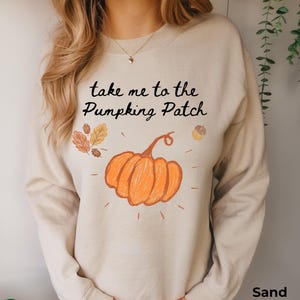 Könnte beinhalten: Ein sandfarbenes Sweatshirt mit Rundhalsausschnitt und dem schwarzen Text "take me to the Pumpking Patch" über einer Grafik eines orangefarbenen Kürbisses, einer kleinen Eichel und herbstlichen Blättern in Braun- und Orangetönen mit Strichen.