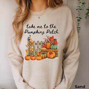 Könnte beinhalten: Ein sandfarbenes Langarm-Sweatshirt mit einem Grafikdruck, der den Text "take me to the Pumpking Patch" über einem Holzzaun, einem Heuballen, verschiedenen orangefarbenen und grünen Kürbissen, Herbstblättern und einem kleinen Vogel auf dem Zaun zeigt.
