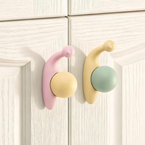 Pode incluir: Dois puxadores de armário em cores pastel. Um é rosa com uma esfera amarela, o outro é amarelo com uma esfera verde. Os puxadores estão montados numa porta de armário branca.