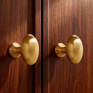 Op de afbeelding: Close-up van twee ovale messing kastknoppen gemonteerd op een donkerbruine houten kastdeur. De knoppen hebben een gladde, gepolijste afwerking en zijn symmetrisch op de deur geplaatst.