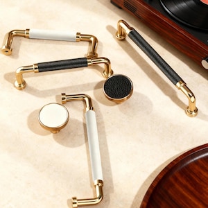 Puede incluir: Una colección de herrajes dorados para armarios, que incluyen tiradores y pomos. Los tiradores presentan empuñaduras envueltas en cuero blanco y negro. Los pomos tienen detalles a juego de cuero y blanco. Los herrajes están dispuestos sobre una superficie clara.