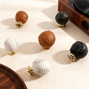 May include: A collection of baseball-themed cabinet knobs. The knobs are made of leather and feature baseball stitching. Colors include brown, black, and white. Each knob has a gold-colored base. The knobs are designed for furniture and home decor.