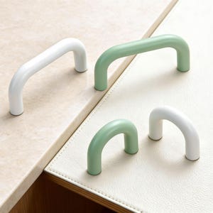 May include: Four cabinet pulls in a minimalist style. Two are white, and two are a soft green. The pulls are arched and have a smooth, rounded design. They are arranged on a light-colored surface.