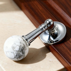 Può includere: Una maniglia per mobili in cromo e marmo. La maniglia presenta uno stelo e una piastra di montaggio in cromo lucido. L'impugnatura è una sfera di marmo bianco con venature grigie. L'hardware è posizionato su una superficie di legno scuro.